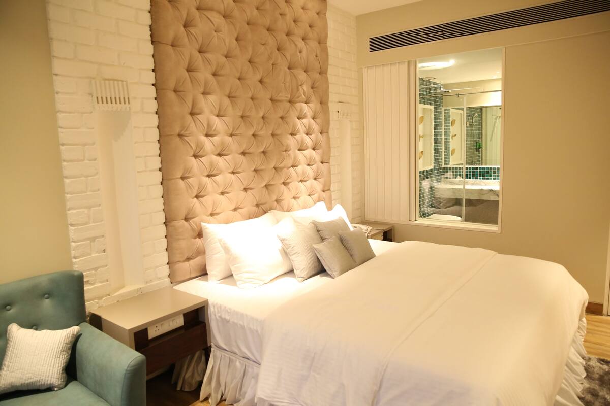 Un dormitorio con un cabecero capitonado, una cama grande, una mesita de noche, un sillón azul y una ventana con vistas al baño.