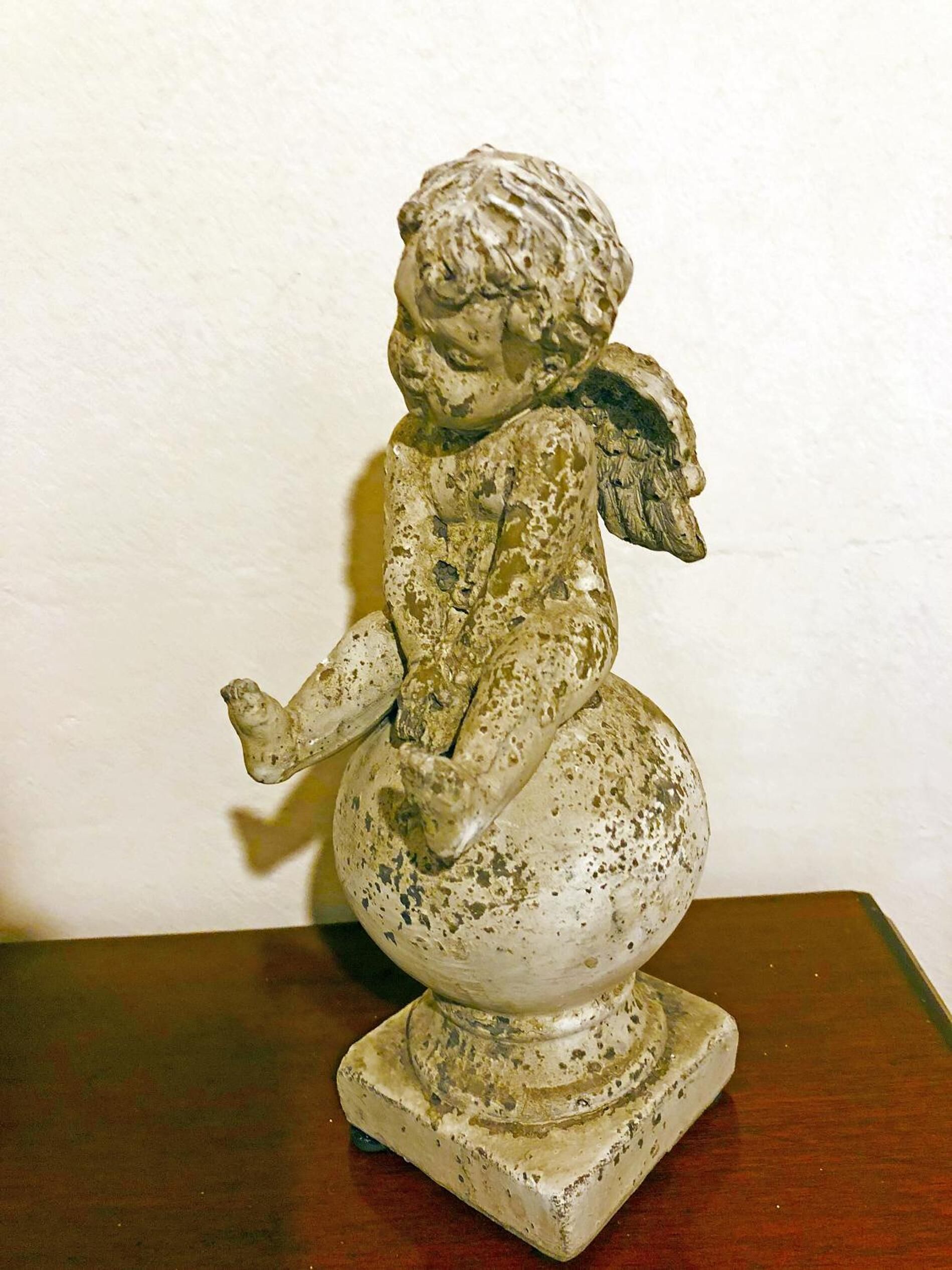 Una pequeña estatua desgastada de una figura sentada sobre un pedestal, ubicada sobre una superficie de madera frente a una pared lisa.