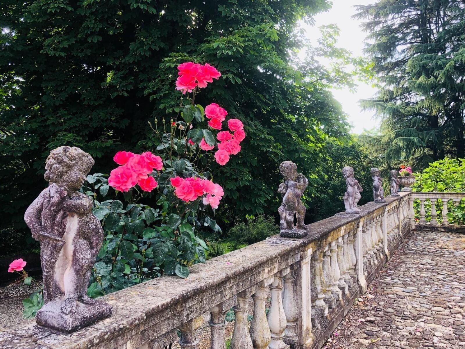 A stone balustrade with statues of cherubs and blooming pink roses in a garden setting.