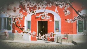 Un bâtiment rouge avec une porte noire, des arches blanches et des volets verts. Il y a deux chaises à l’extérieur.