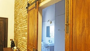 Une pièce avec un mur en briques, des portes en bois, une petite table avec une machine à café et des pâtisseries, et une salle de bain visible à travers une porte ouverte.
