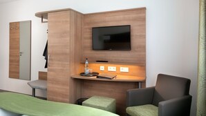 A modern hotel room with a wooden accent wall, a flat-screen TV mounted above a desk, a mirror, a green bed, and two armchairs. - Hotel Kern (Walddorfhäslach)