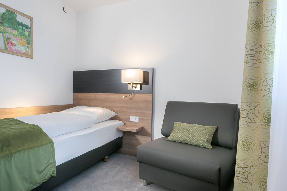 A hotel room with a bed, a nightstand, a lamp, a chair, and a painting on the wall.