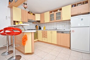 A kitchen with wooden cabinets, a white refrigerator, and two red bar stools. - Apartments Miroslava (Vodice)