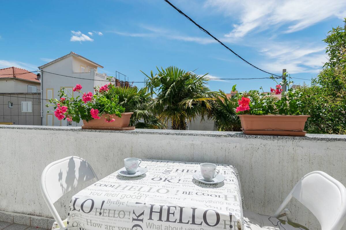 A terrace with a table set for two, surrounded by potted plants and a clear blue sky.