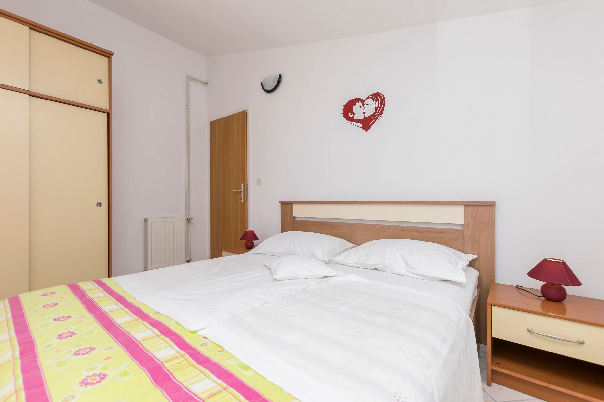 A neatly made bed with white bedding, a wooden headboard, a bedside table with a lamp, and a wall clock.
