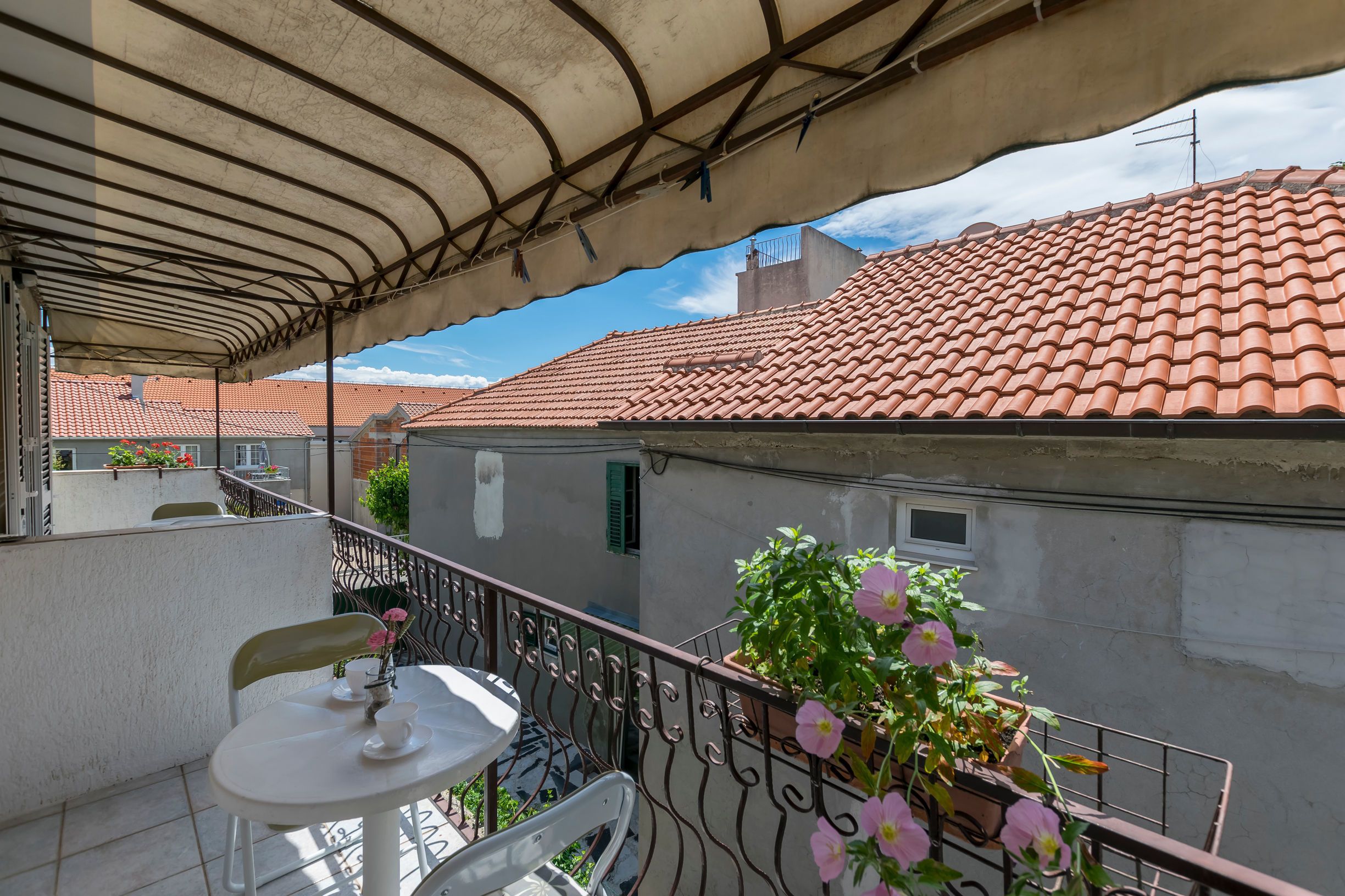 Ein Balkon mit Tisch und Stühlen, ein Blumentopf mit rosa Blüten und ein rotes Ziegeldach im Hintergrund.