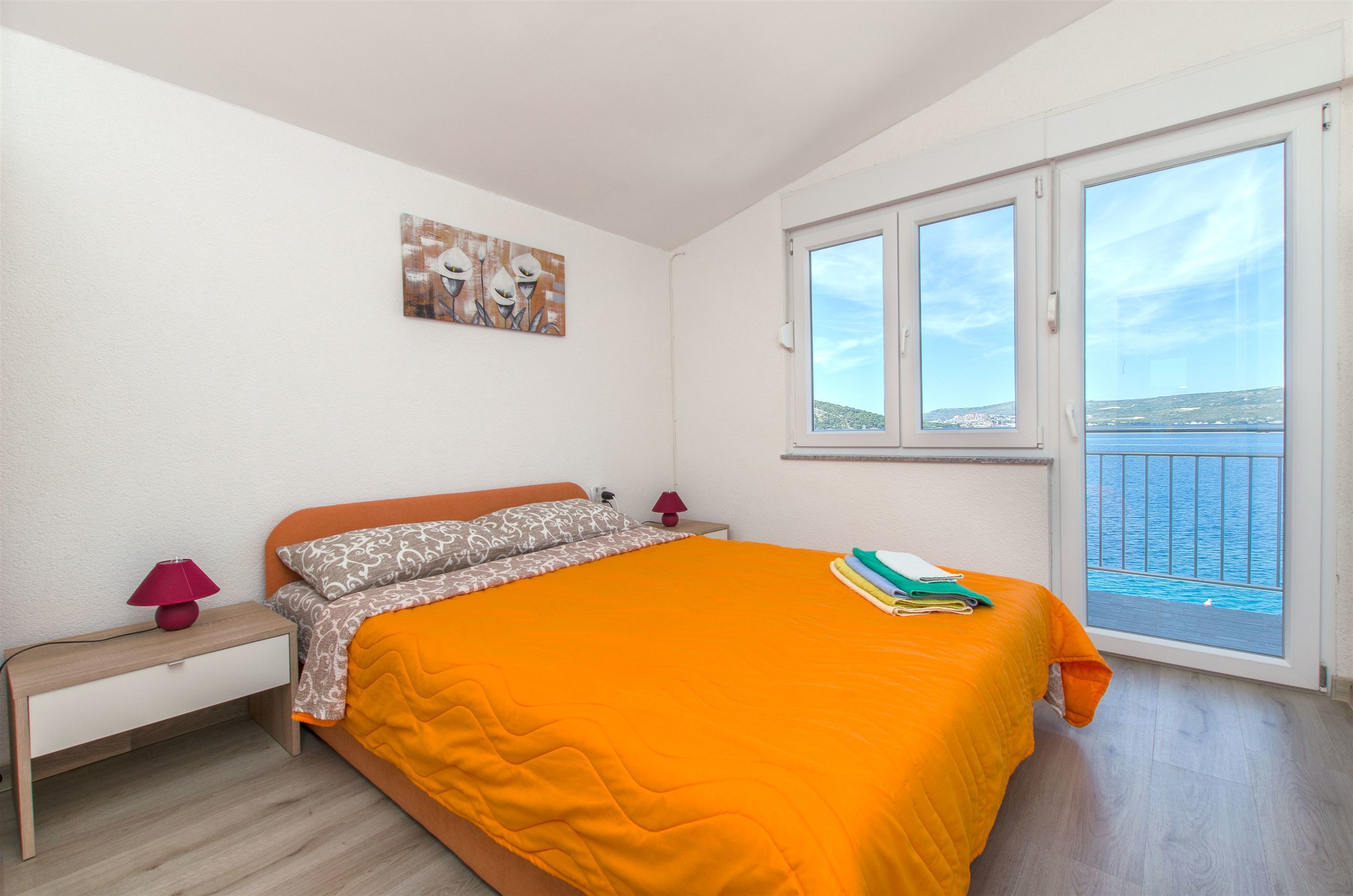 Une chambre à coucher avec un lit, une table de chevet, une lampe, un tableau et une fenêtre donnant sur la mer.