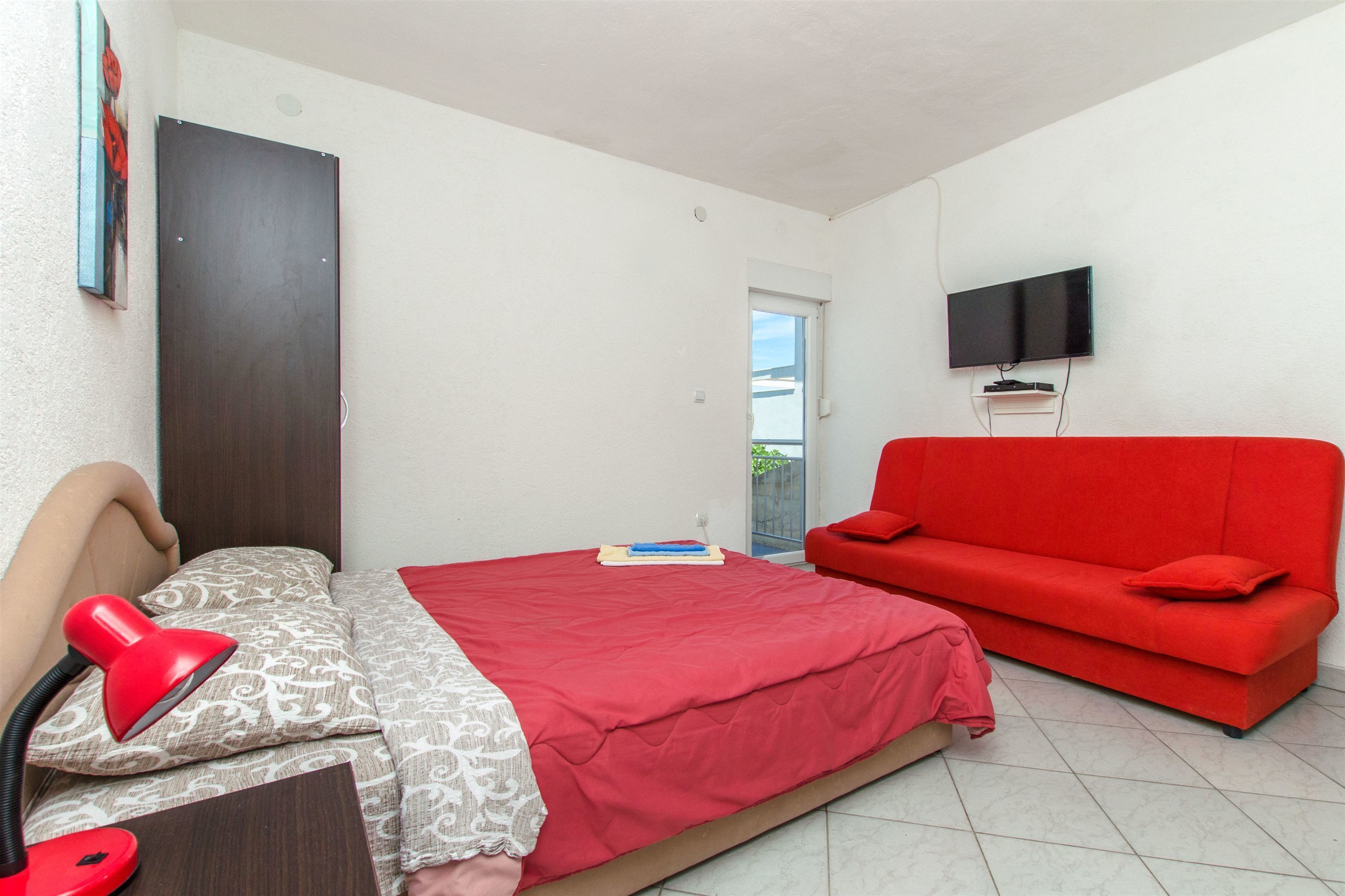 A bedroom with a bed, a red sofa, a TV mounted on the wall, and a door leading to a balcony.