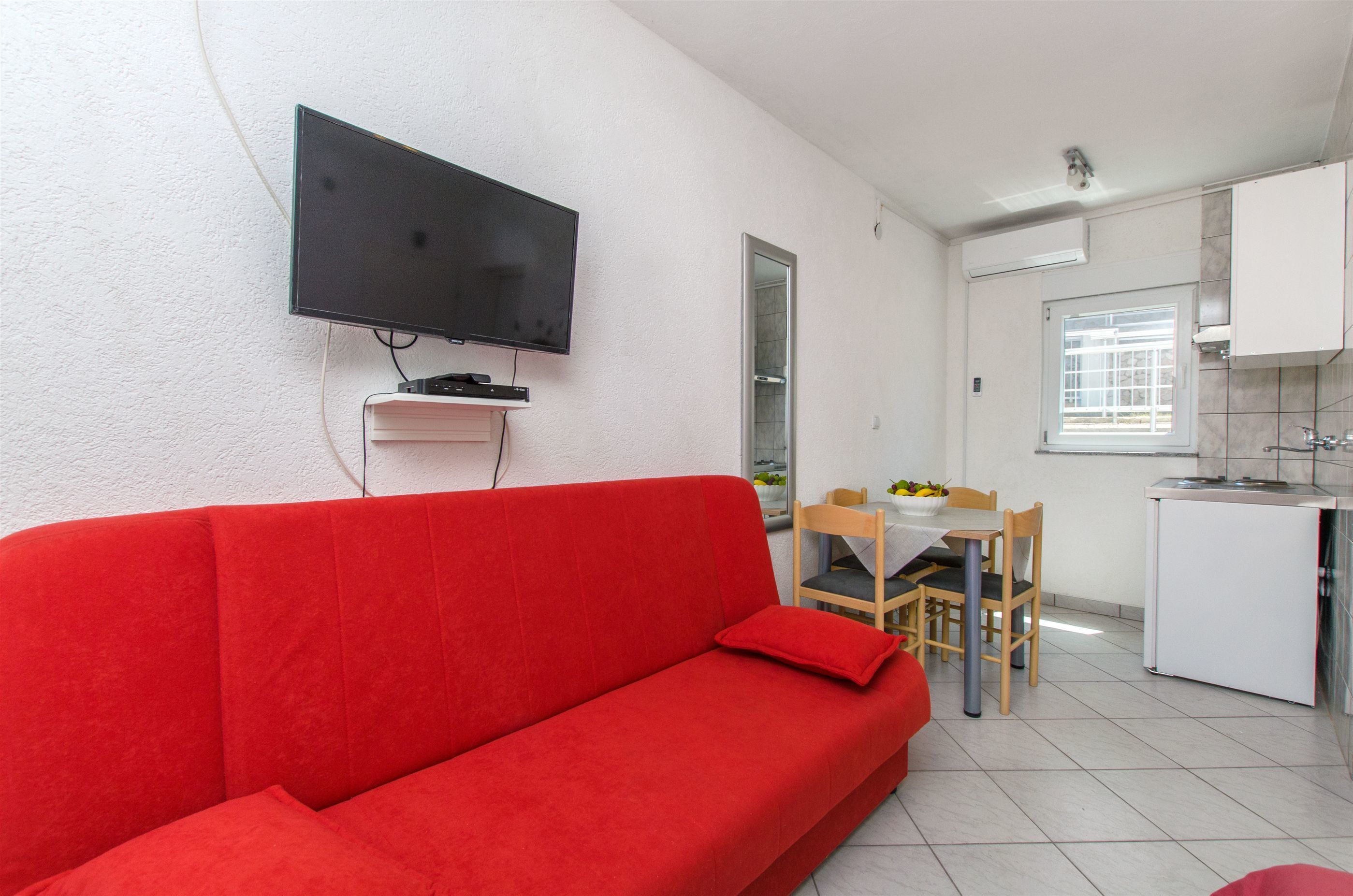 Ein rotes Sofa in einem Wohnbereich mit einem an der Wand montierten Fernseher, einem Esstisch mit Stühlen sowie einer Küchenzeile mit Schränken und einem Kühlschrank.