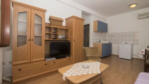 A compact living space with a wooden cabinet, a television, a small table with a lace tablecloth, and a kitchenette area. - Apartments Jaksa (Jelsa)