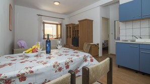 A dining area with a table set for two, a fruit bowl, wine glasses, and a bottle. There is a window with curtains, a wooden cabinet, and a kitchen area with blue cabinets. - Apartments Jaksa (Jelsa)