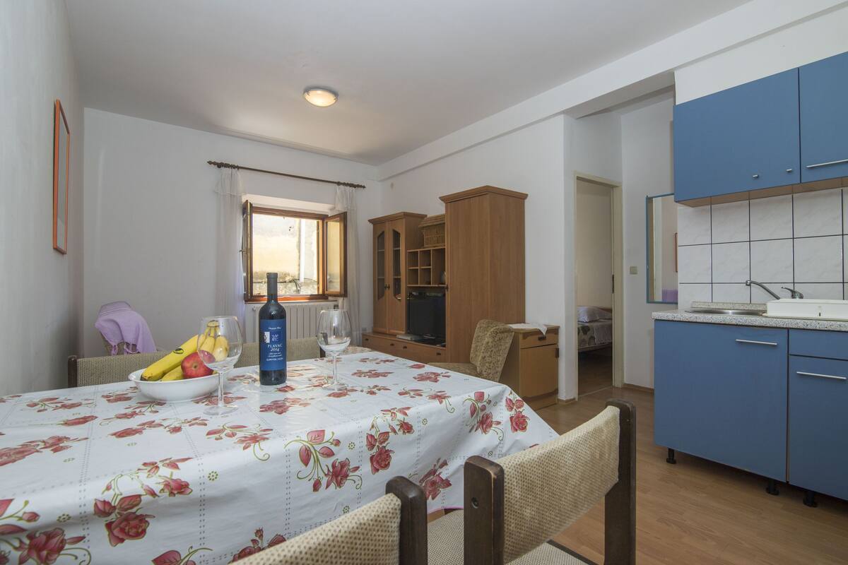 A dining area with a table set for two, a kitchen with blue cabinets, and a window with curtains.