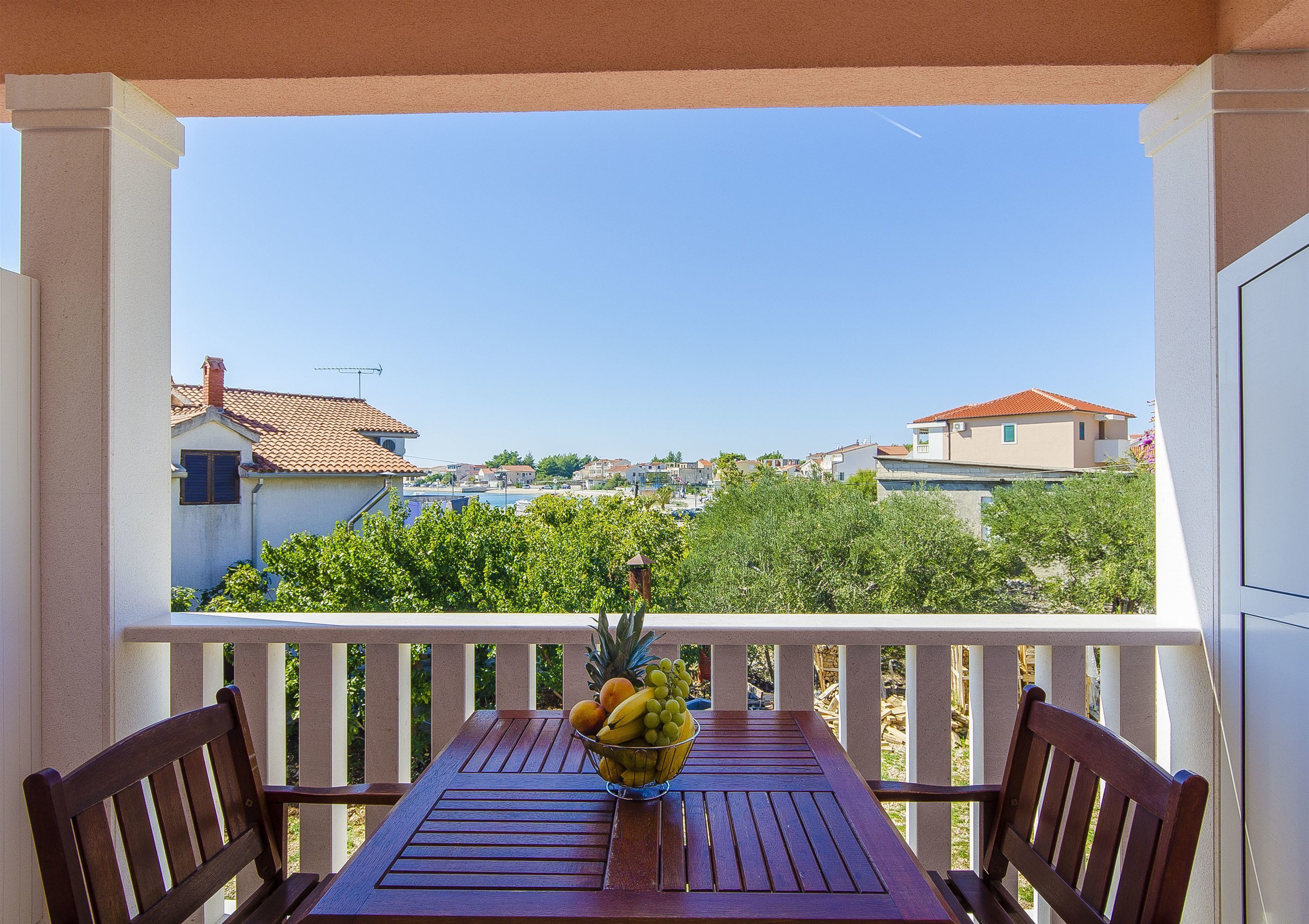 Ein Balkon mit Holztisch und Stühlen, einer Obstschale und Blick auf Häuser und Bäume.