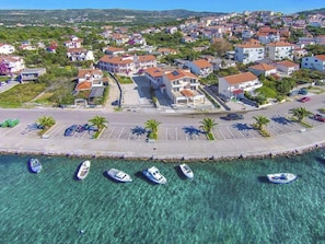 Vista aerea di una cittadina costiera con acque turchesi cristalline, un porto turistico con diverse imbarcazioni e una varietà di edifici residenziali.