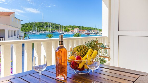 Un balcon avec une table en bois, une bouteille de vin rosé, un panier de fruits et deux verres à vin.