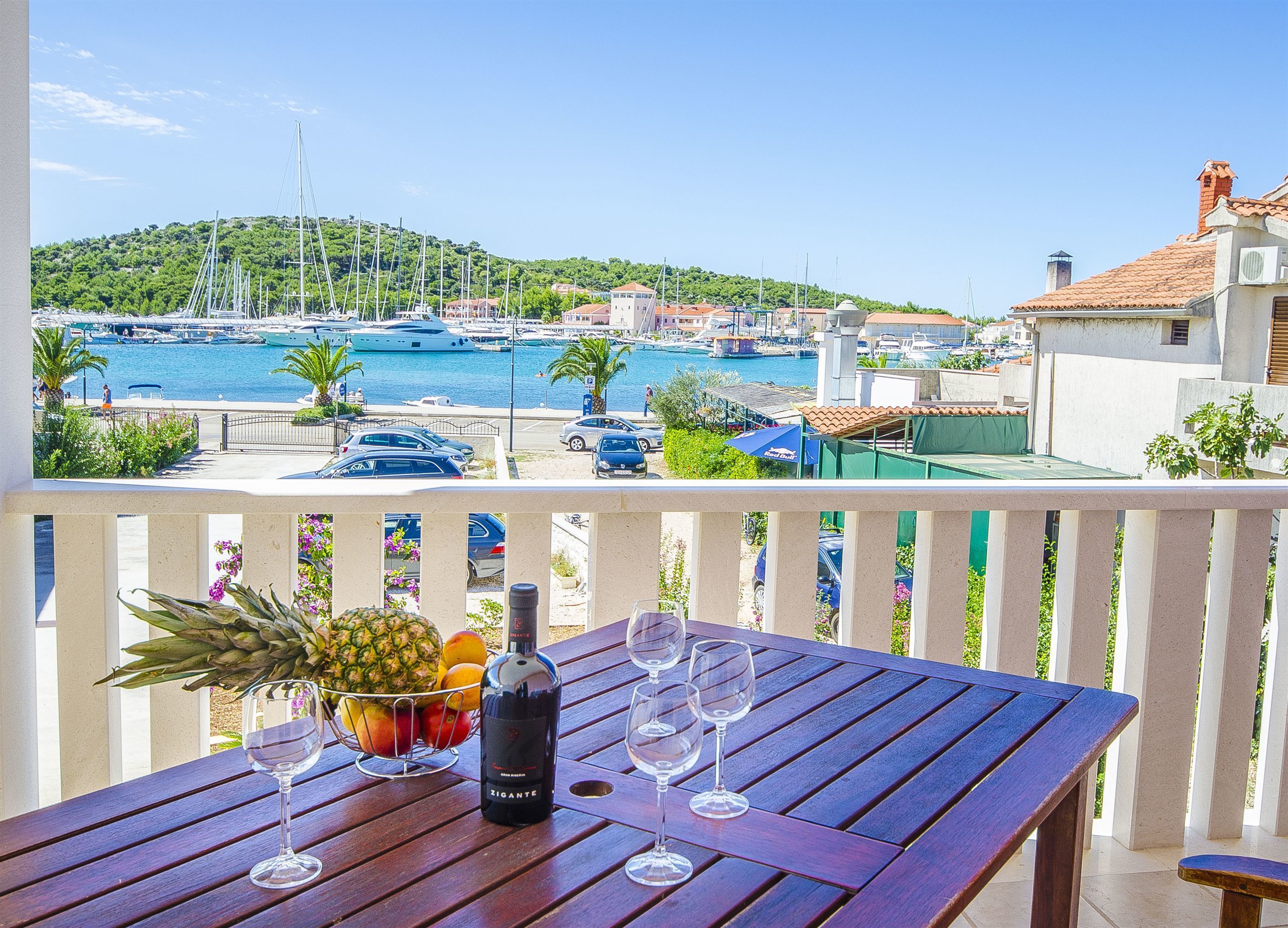 Et træbord med en flaske vin, glas og en frugtkurv på en balkon med udsigt over en havn fyldt med både og bygninger.