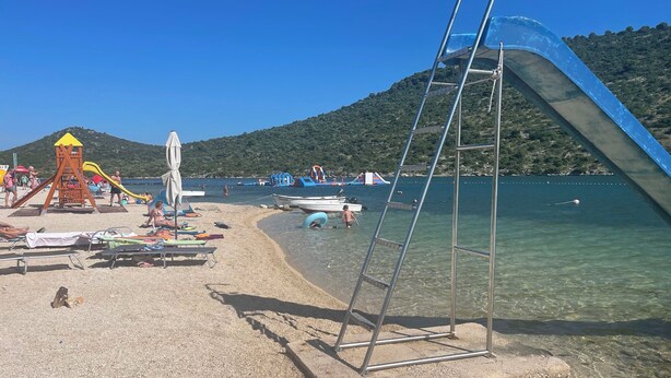 Una playa con un parque infantil, agua cristalina y barcos.