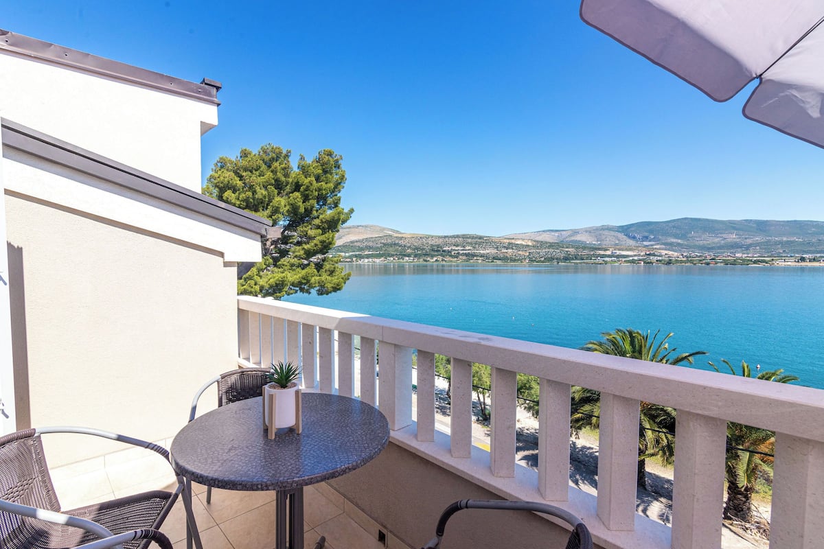 A balcony with a round table and chairs, overlooking a clear blue sea and distant hills.