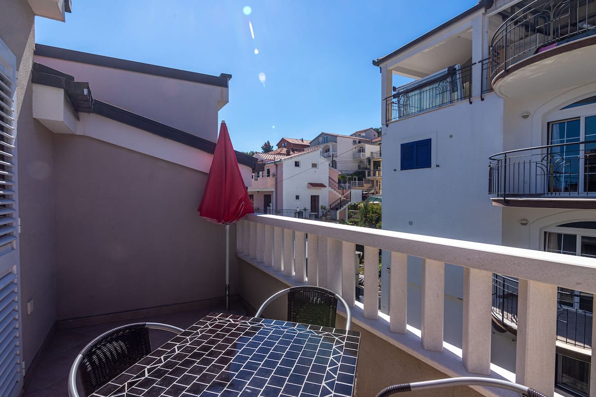 A balcony with a tiled table and chairs, a red umbrella, and a view of neighboring buildings.