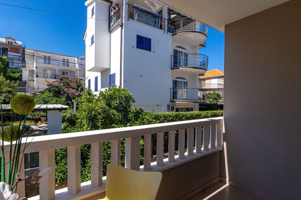 A balcony with a white railing, overlooking a residential area with white buildings and green trees.