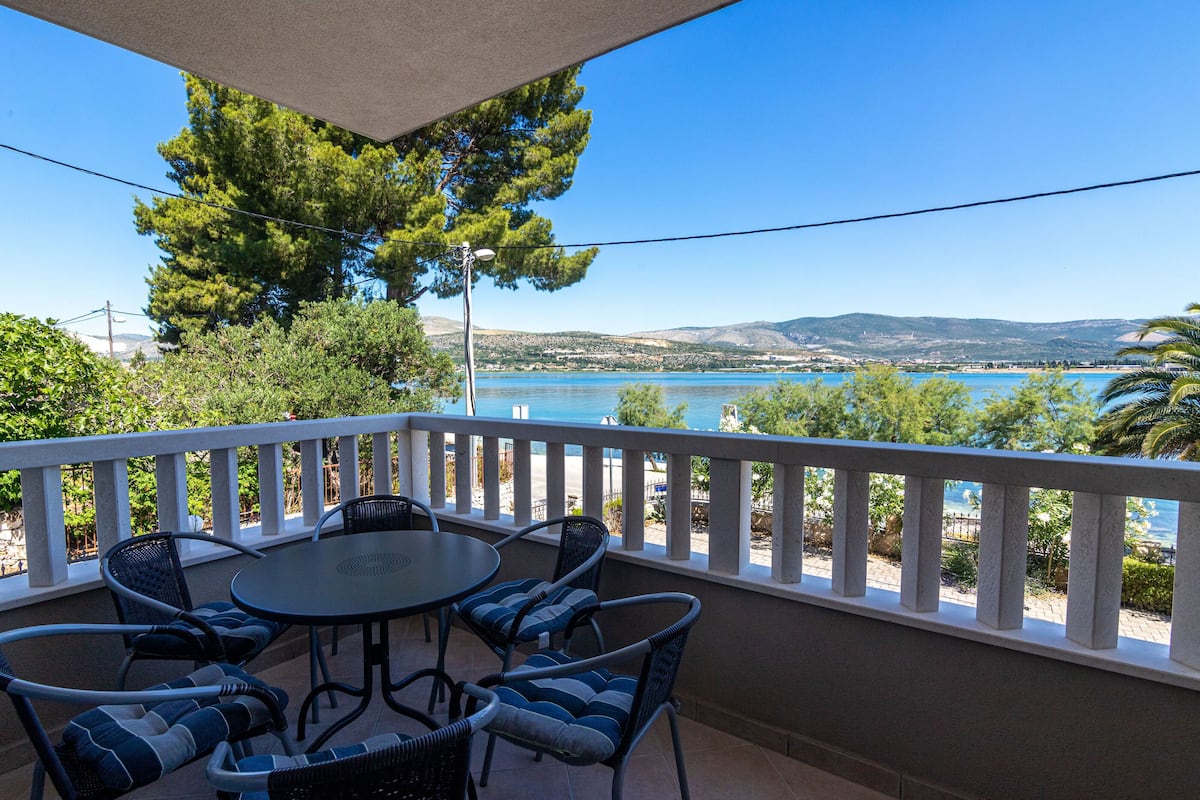 A balcony with a table and chairs, overlooking a body of water and a hilly landscape.