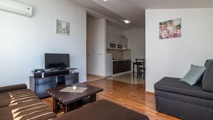 Un salon moderne avec un canapé, une table basse et un téléviseur. On aperçoit une cuisine à l’arrière-plan, avec des placards et un four à micro-ondes.