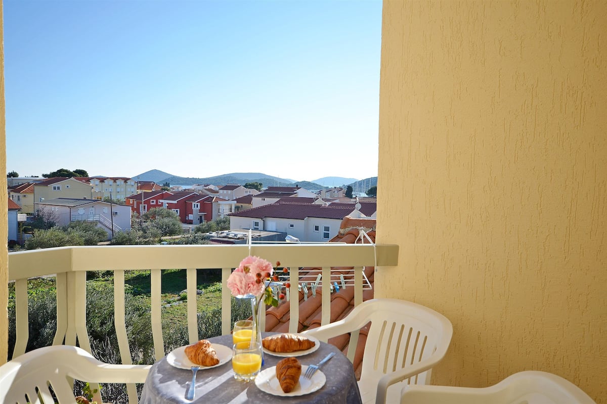 A balcony with a table set for breakfast, including croissants and orange juice, overlooking a residential area with houses and hills.
