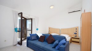 A modern bedroom with a blue sofa, a wooden wardrobe, a bedside table with a lamp, and a large window with curtains. - Apartments Mira (Tribunj)