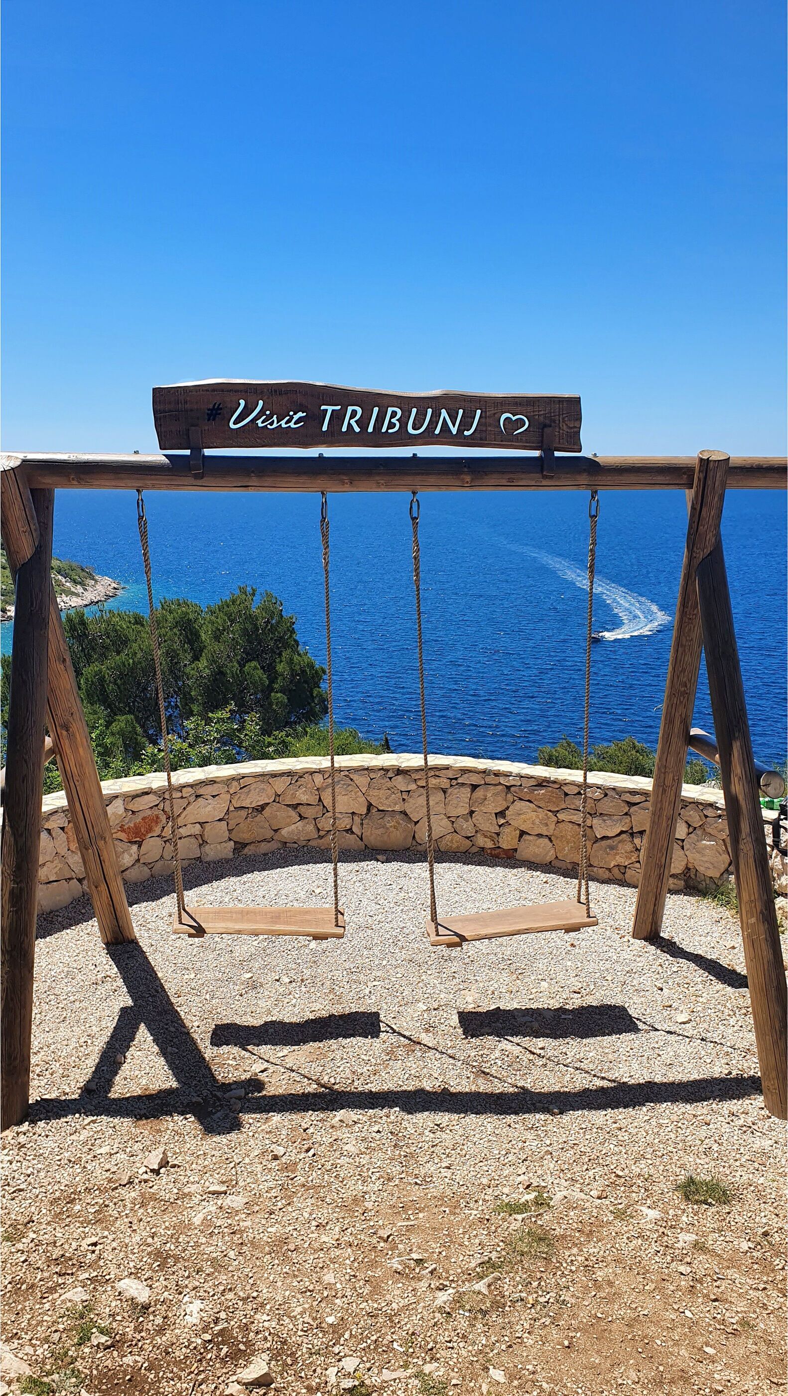 Un’altalena in legno con un cartello che recita 'Visit TRIBUNJ' sullo sfondo di un cielo azzurro e vista sul mare.