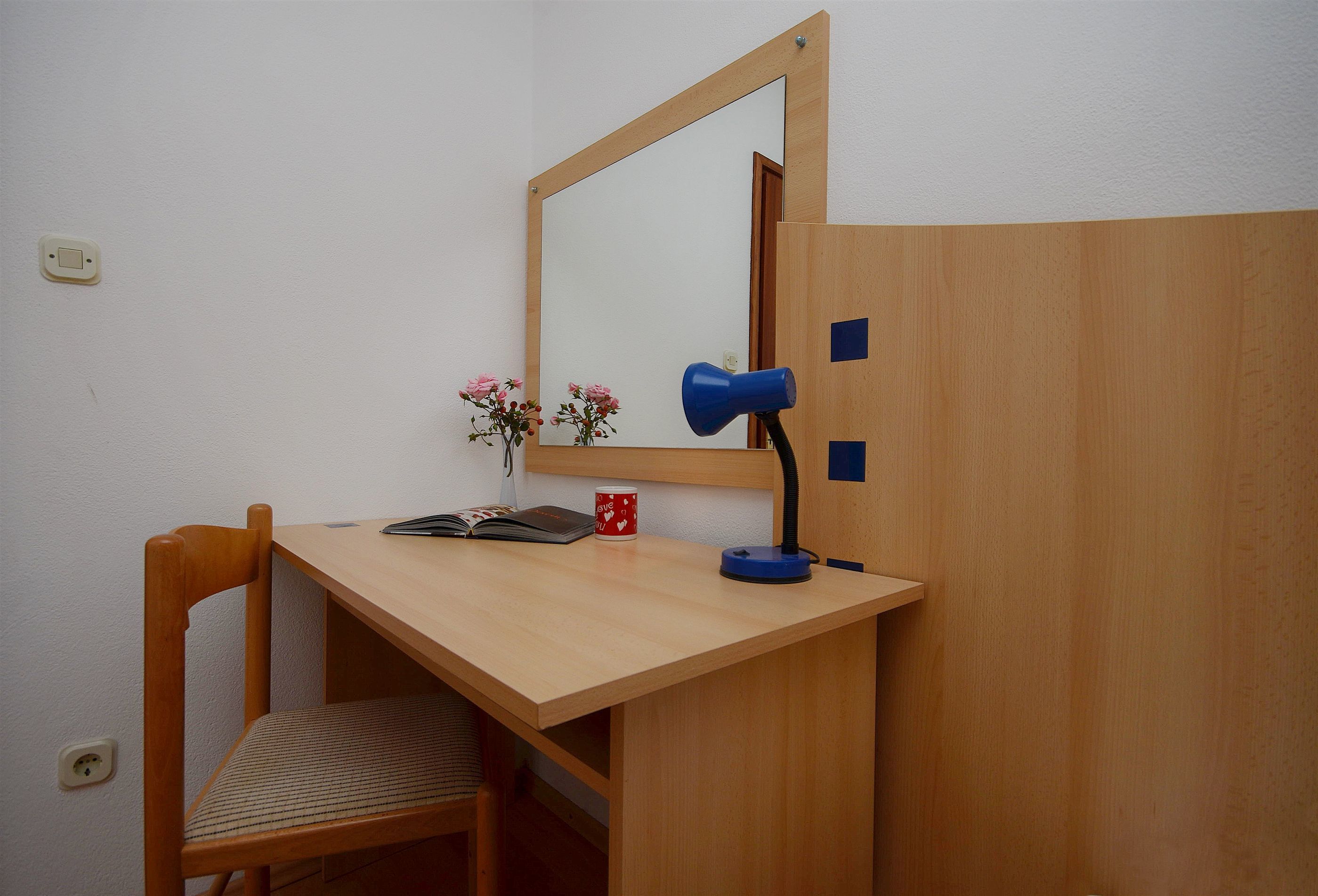 A wooden desk with a mirror, a blue lamp, a red mug, and a book.
