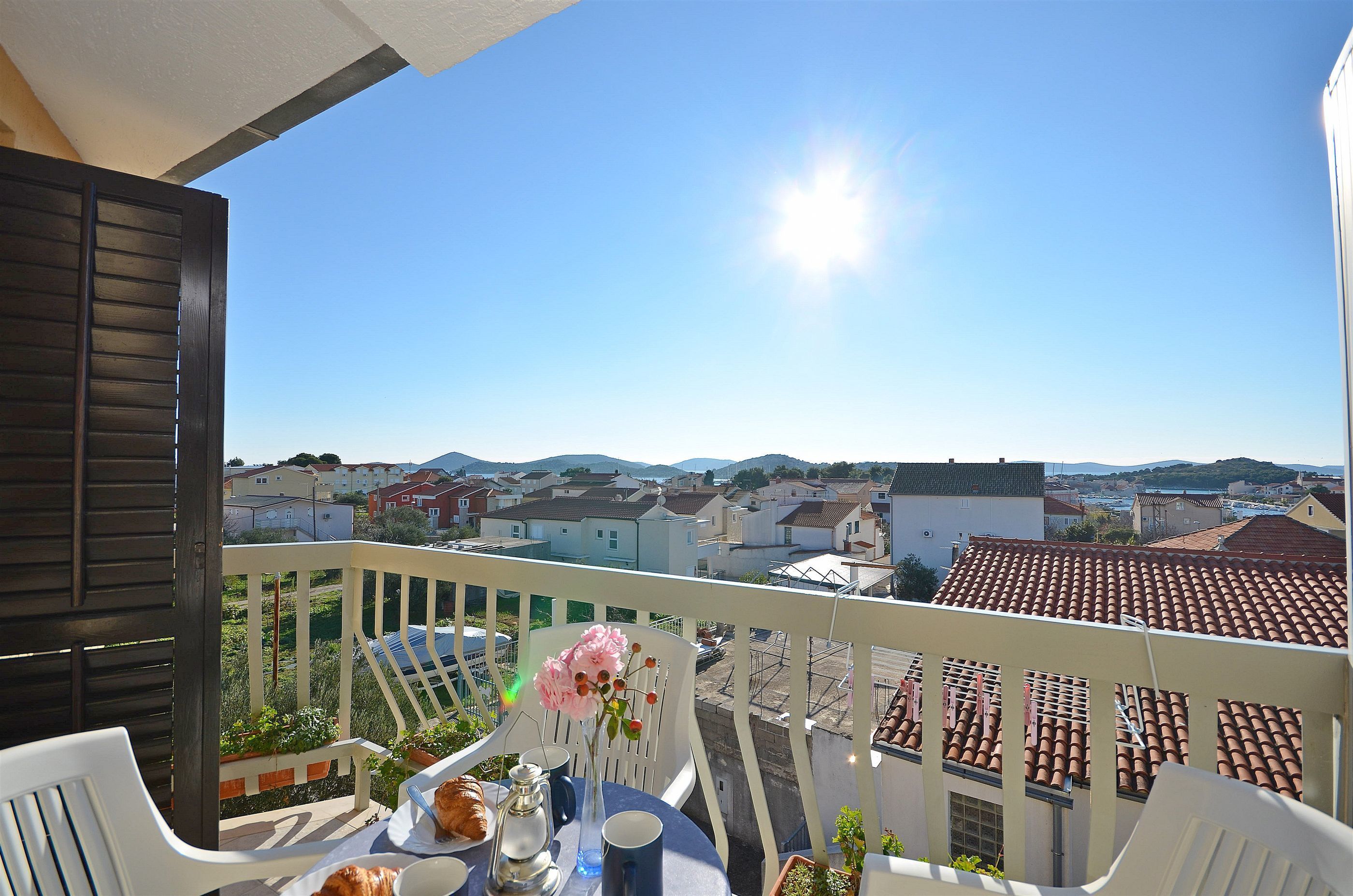 En balkon med et bord dækket til to, med udsigt over et boligområde med huse og en klar blå himmel.