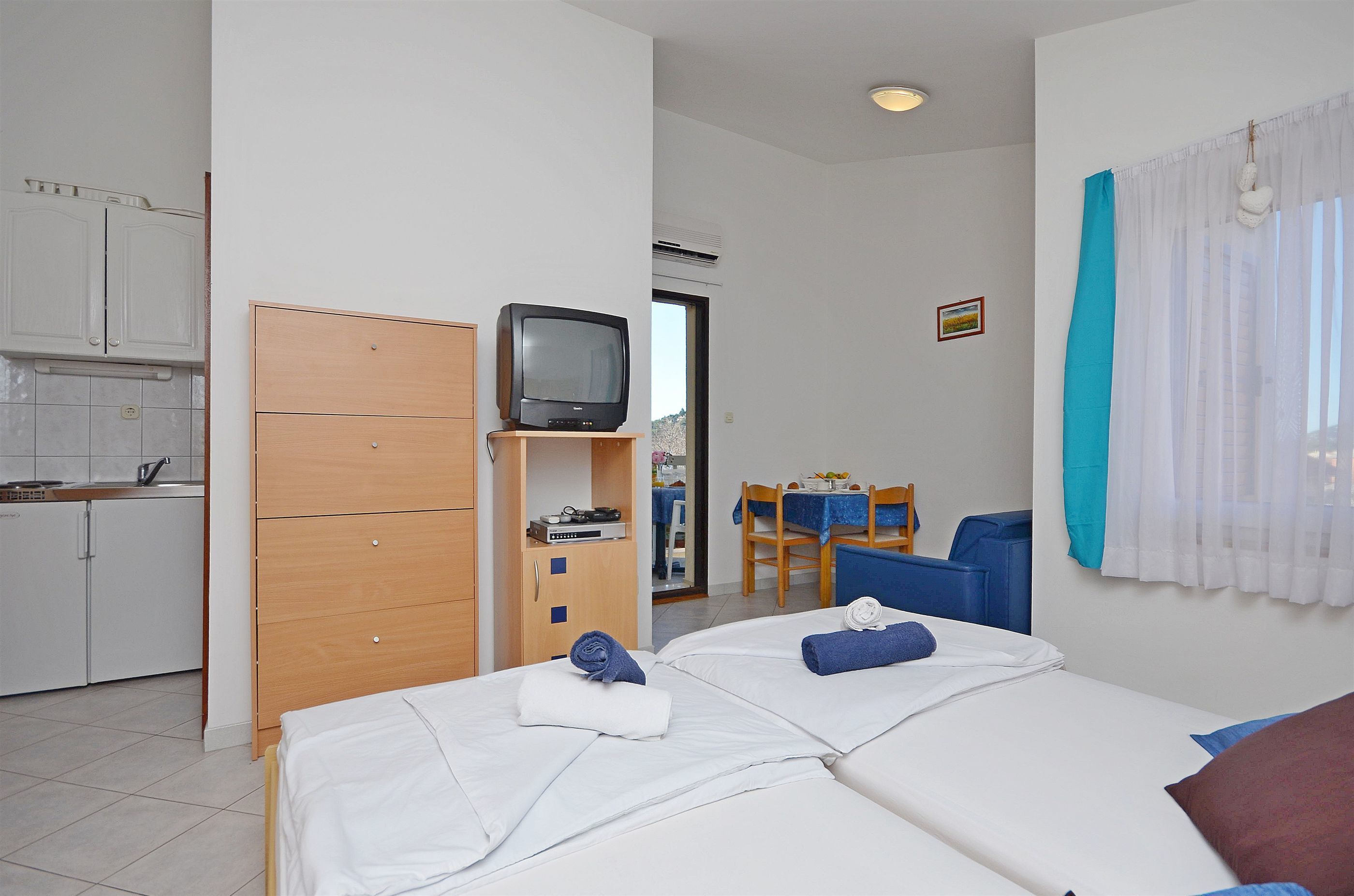 A hotel room with two beds, a wooden wardrobe, a television on a stand, a dining area with a table and chairs, and a bathroom visible through an open door.