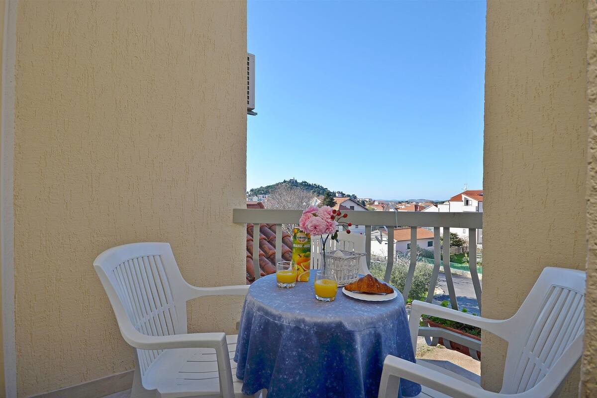 A balcony with a table set for breakfast, including orange juice, a croissant, and a vase with flowers.