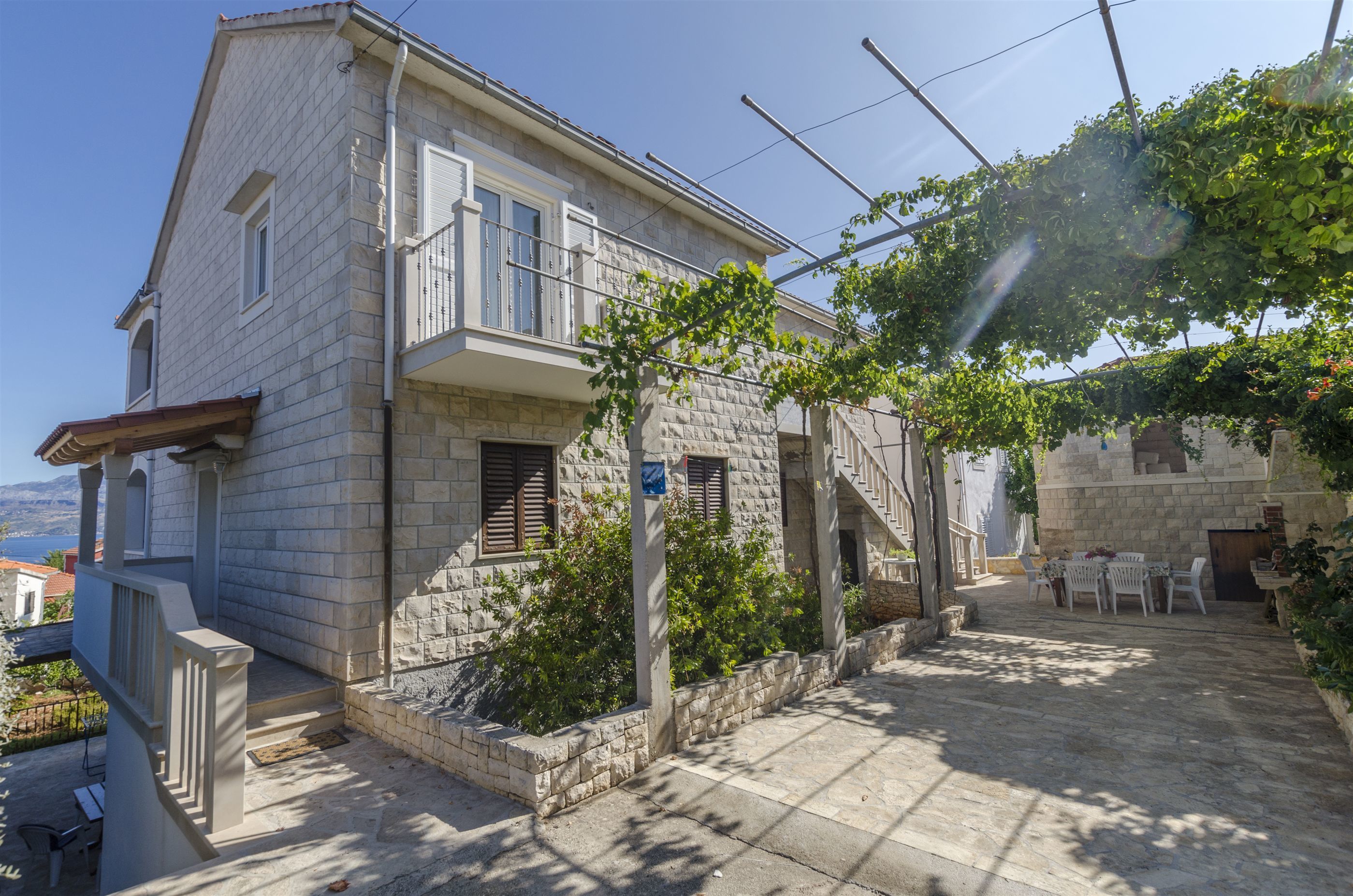 A stone house with a balcony, a garden, and a table with chairs outside.