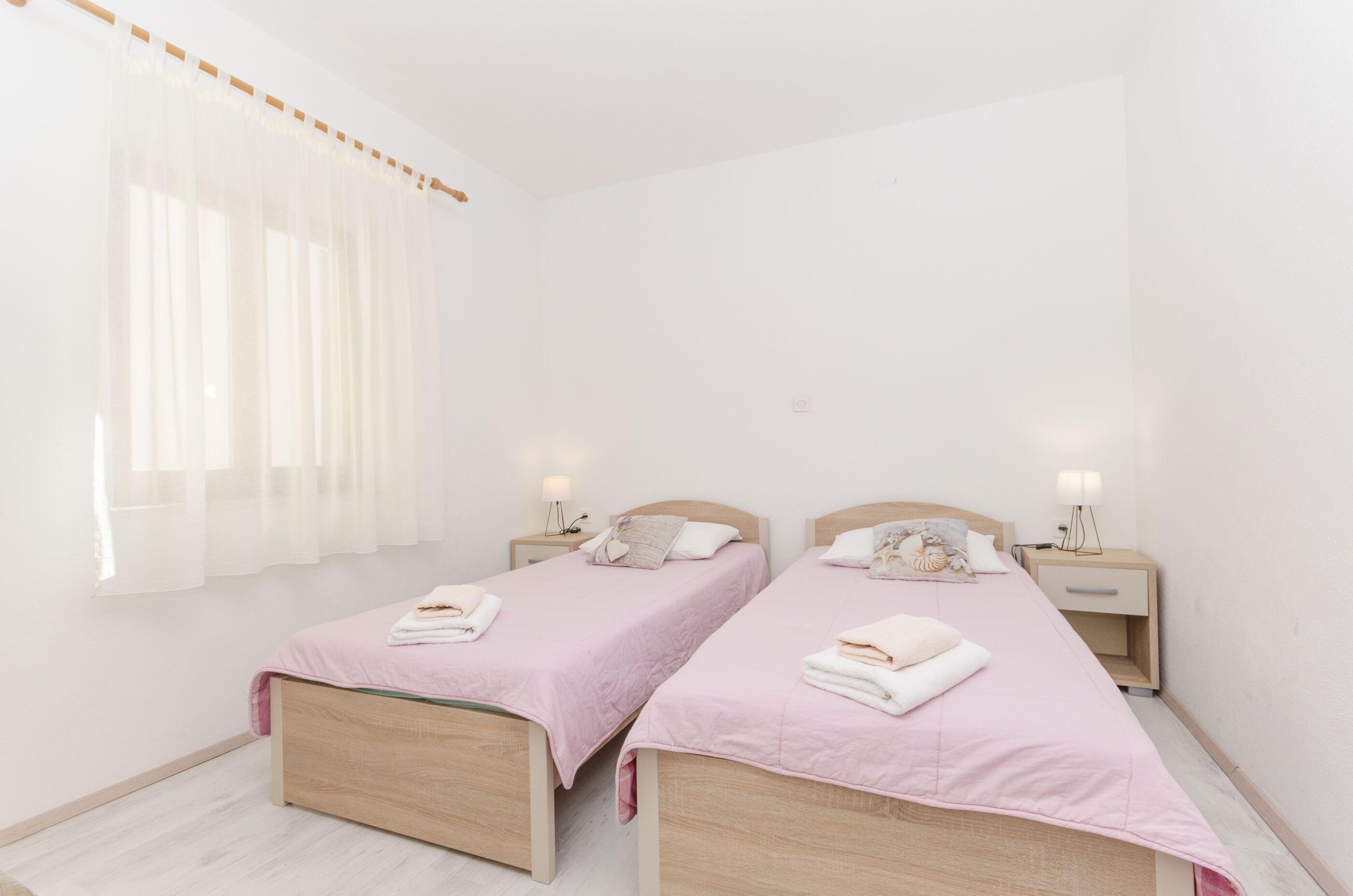 Ein Schlafzimmer mit zwei Betten, beide mit Holzrahmen und rosa Bettwäsche. Es gibt Nachttische mit Lampen und ein Fenster mit durchsichtigen Vorhängen.