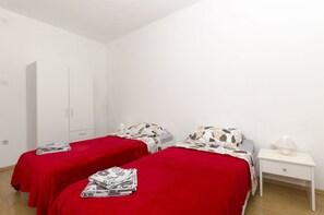 Ein Zimmer mit zwei Einzelbetten, jeweils mit roter Bettdecke und einem gemusterten Kopfkissen. Ein weißer Nachttisch mit Lampe steht daneben, und in der Ecke ist ein weißer Kleiderschrank.