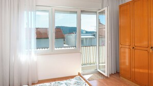 A room with a large window, wooden wardrobe, and a view of a balcony with a tiled roof. - Apartment Luketa (Primosten)