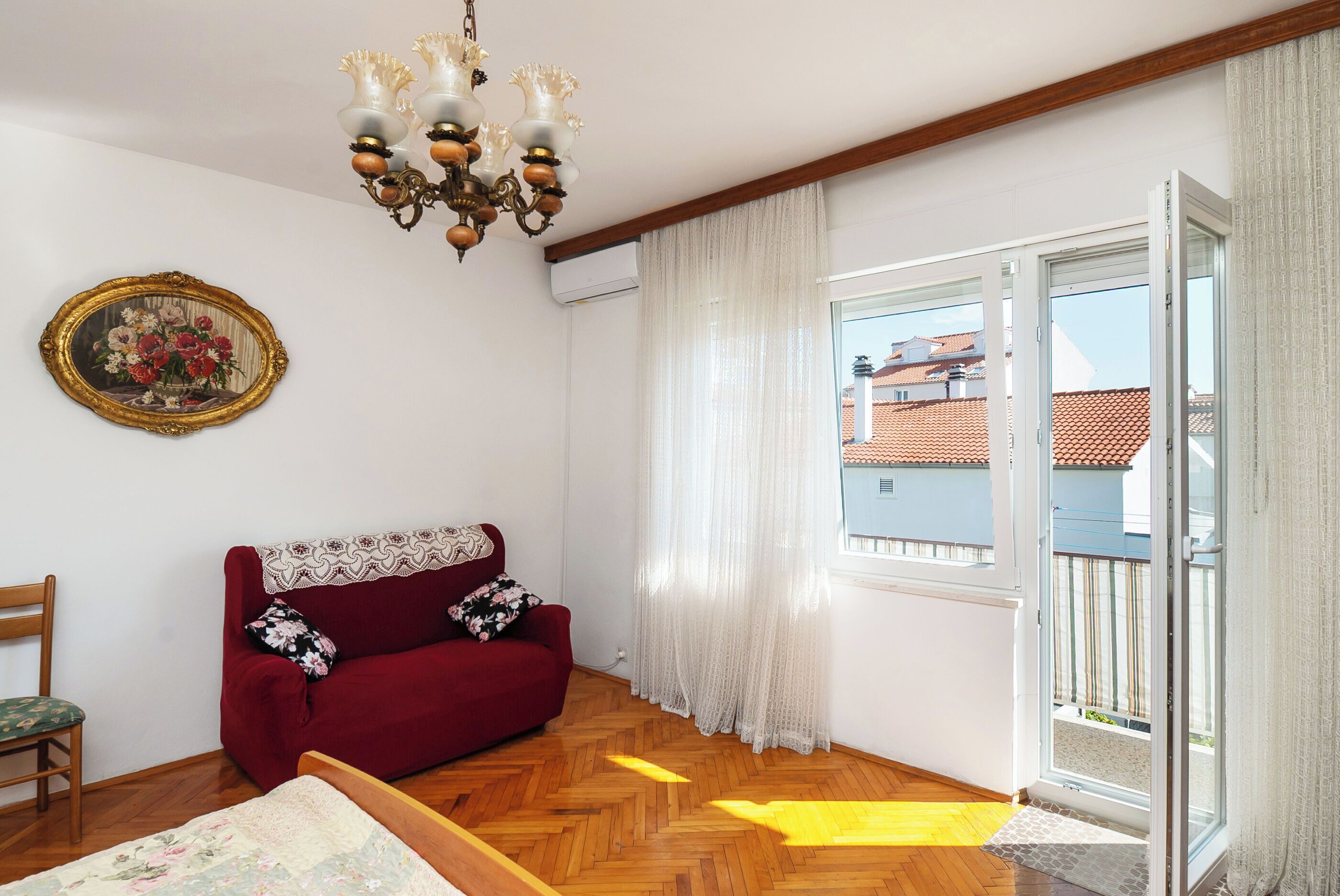 Una habitación con un sofá rojo, piso de madera, una araña, una ventana con cortinas y un cuadro en la pared.