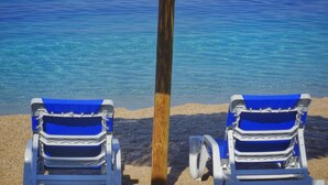 Two beach chairs under a thatched umbrella on a sandy beach with clear blue water. - Apartments Kure (Primosten)