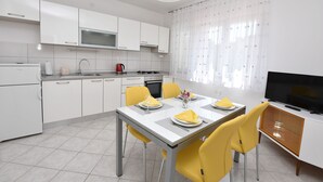 A modern kitchen with white cabinets, a dining table set for four, yellow chairs, and a TV on a wooden cabinet. - Apartments Kure (Primosten)