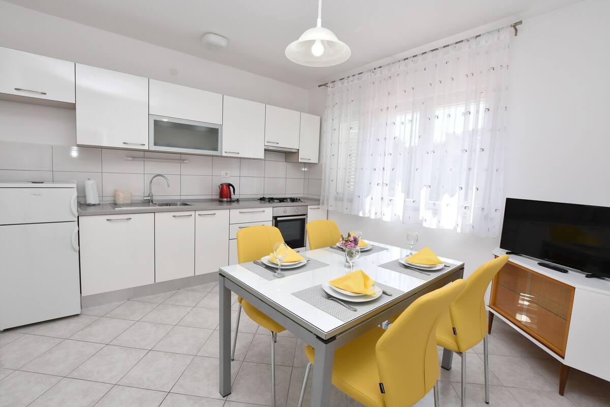 A modern kitchen with white cabinets, a dining table set for four, yellow chairs, and a TV on a wooden cabinet.