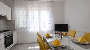 A dining area with a table set for four, yellow chairs, a flat-screen TV, and a kitchen with white cabinets and a stove. - Apartments Kure (Primosten)