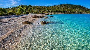 A clear beach with pebbles and rocks, a calm sea, and a green hill in the background. - Apartments Kure (Primosten)