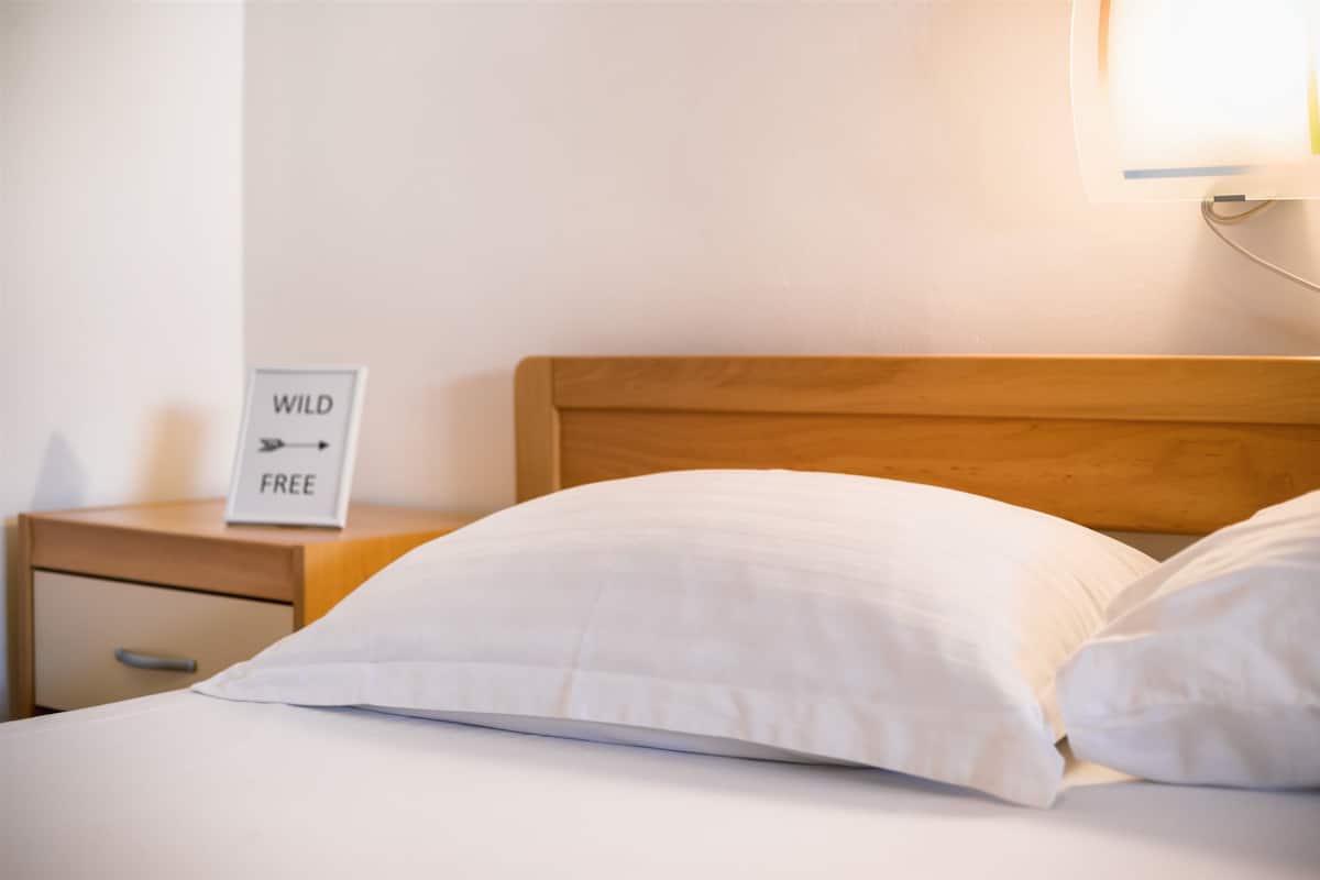 A neatly made bed with white bedding and a wooden headboard. A small bedside table holds a sign that reads "WILD FREE."