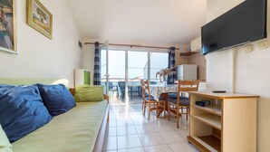 A cozy living room with a sofa, a dining area with chairs and a table, a TV, and a view through the large window. - Apartments Zanze (Vodice)