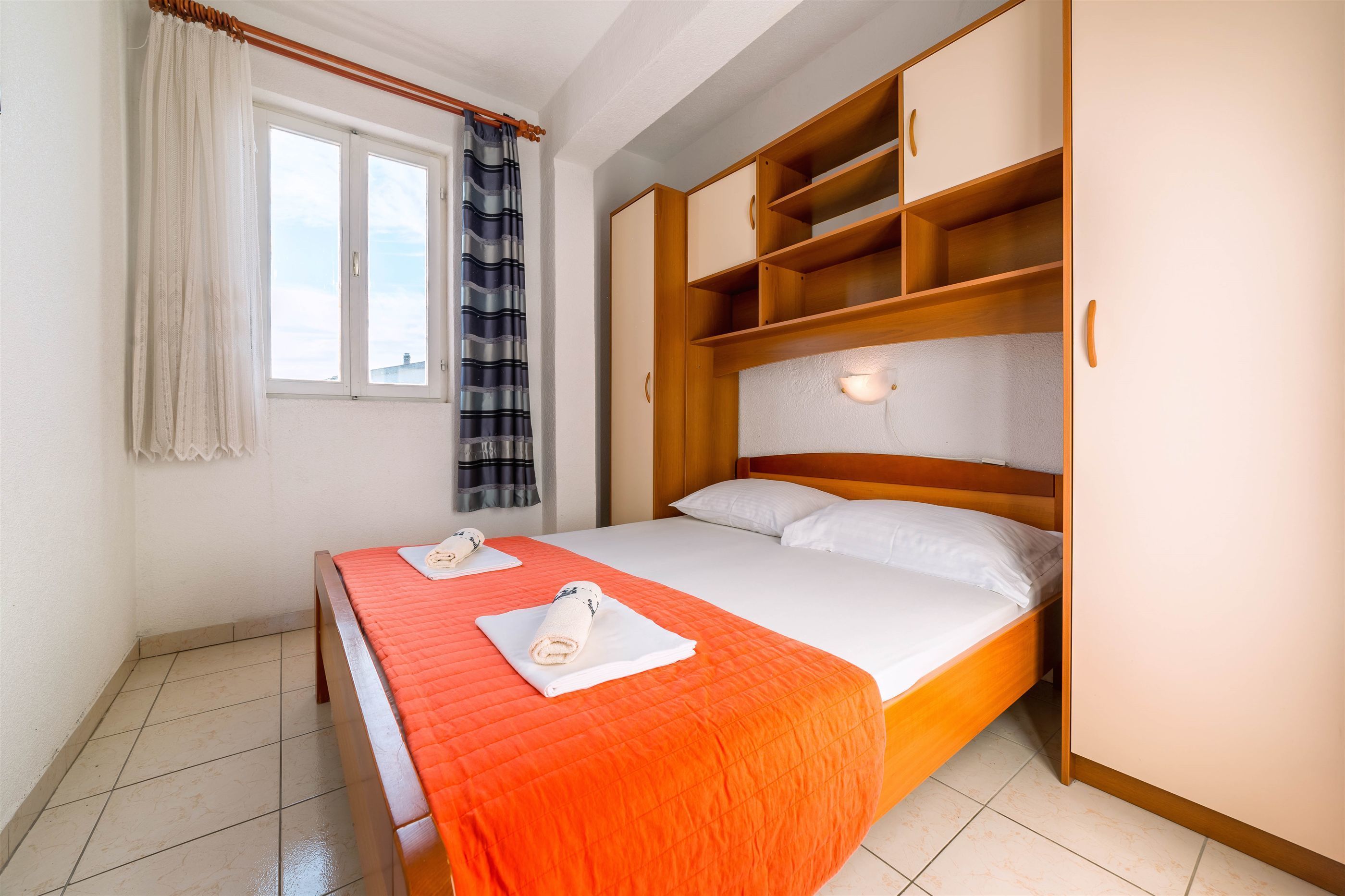 A double bed with an orange bedspread, a wooden wardrobe with multiple compartments, a window with curtains, and a tiled floor.