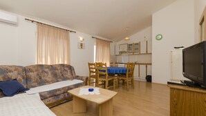 Un salon avec un canapé, une table basse, un téléviseur posé sur un meuble, un coin repas avec une table et des chaises, et une cuisine à l’arrière-plan.