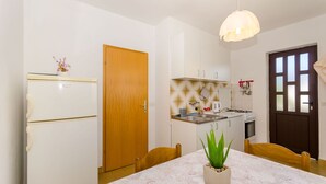 A compact kitchen with white cabinets, a wooden door, a dining table with chairs, and a hanging light fixture. - Apartment Lavcevic (Vrboska)