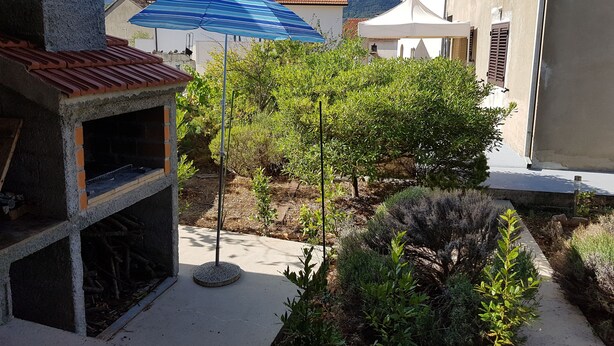 A courtyard with a barbecue area, a blue umbrella, and surrounding greenery. - Apartment Lavcevic (Vrboska)